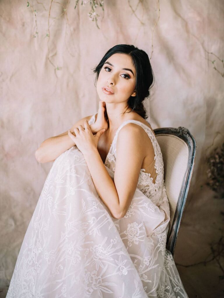 Bride-to-be seated on a vintage chair wearing a lace gown, surrounded by soft pastel hues and hanging greenery in the Vella Nest Floral studio.