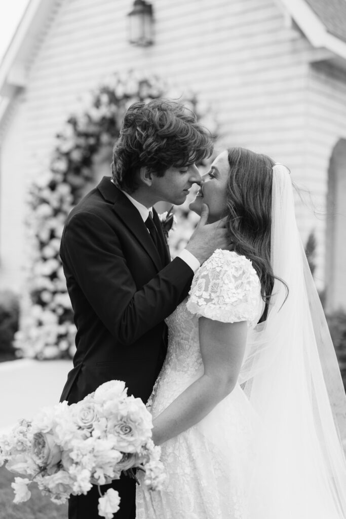 A bride and groom stand beneath a soft, garden‑inspired floral arch, sharing an intimate moment outside a light‑siding chapel during their Dallas–Fort Worth wedding.